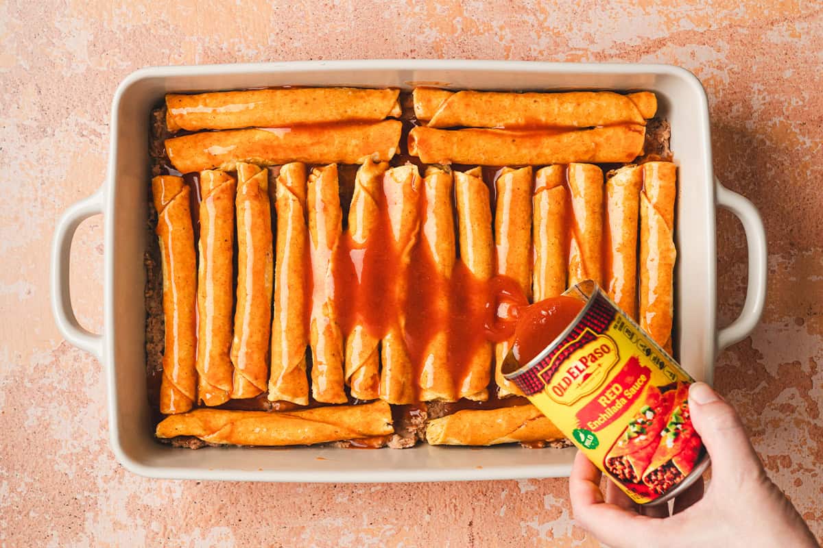 Enchilada sauce being poured over taquitos in a baking dish.
