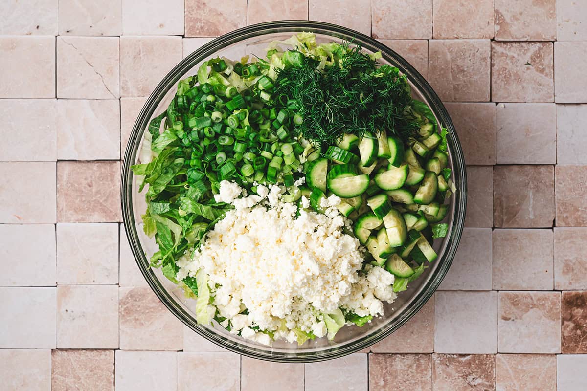 Crumbled feta, sliced cucumber, dill, green onions and lettuce in a bowl.