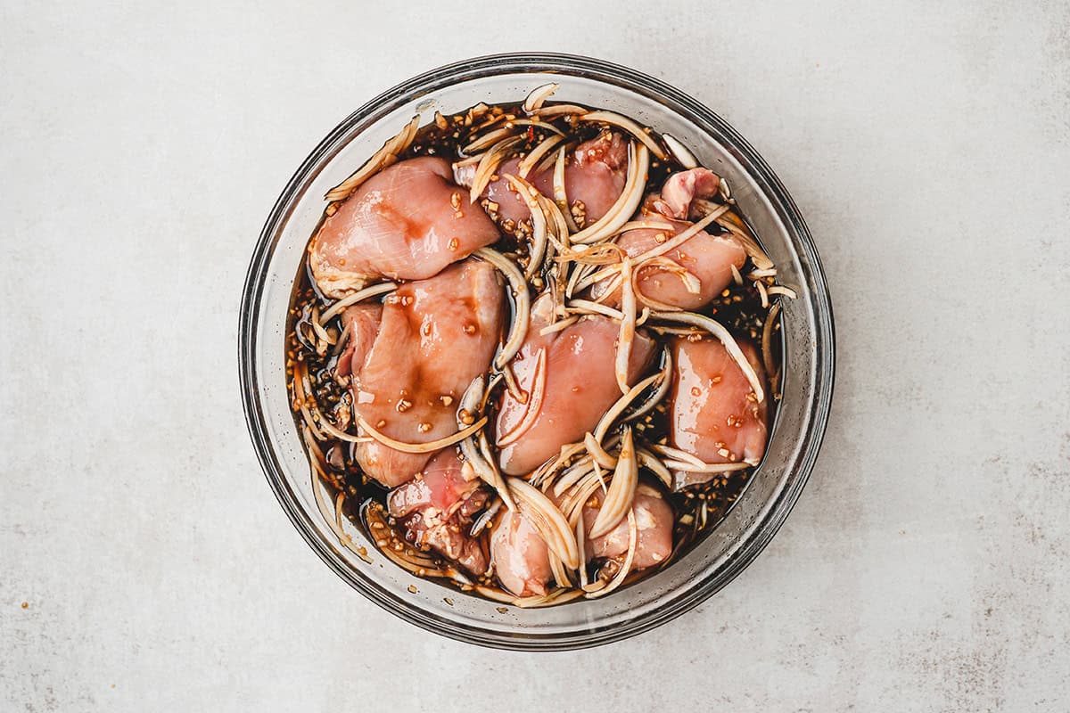 Chicken thighs marinating in pineapple soy sauce marinade with sliced onions in a glass bowl
