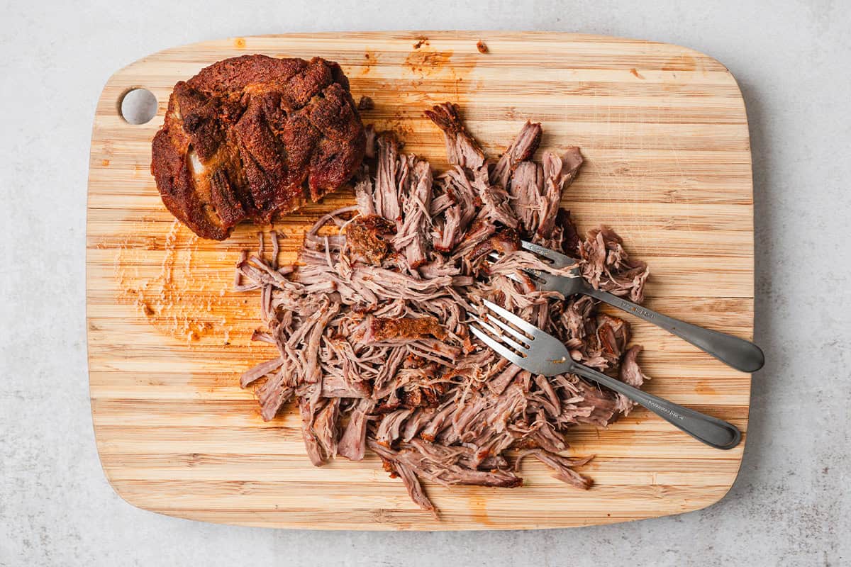 Fork-tender pulled pork being shredded on a wooden cutting board with crispy browned bark pieces.