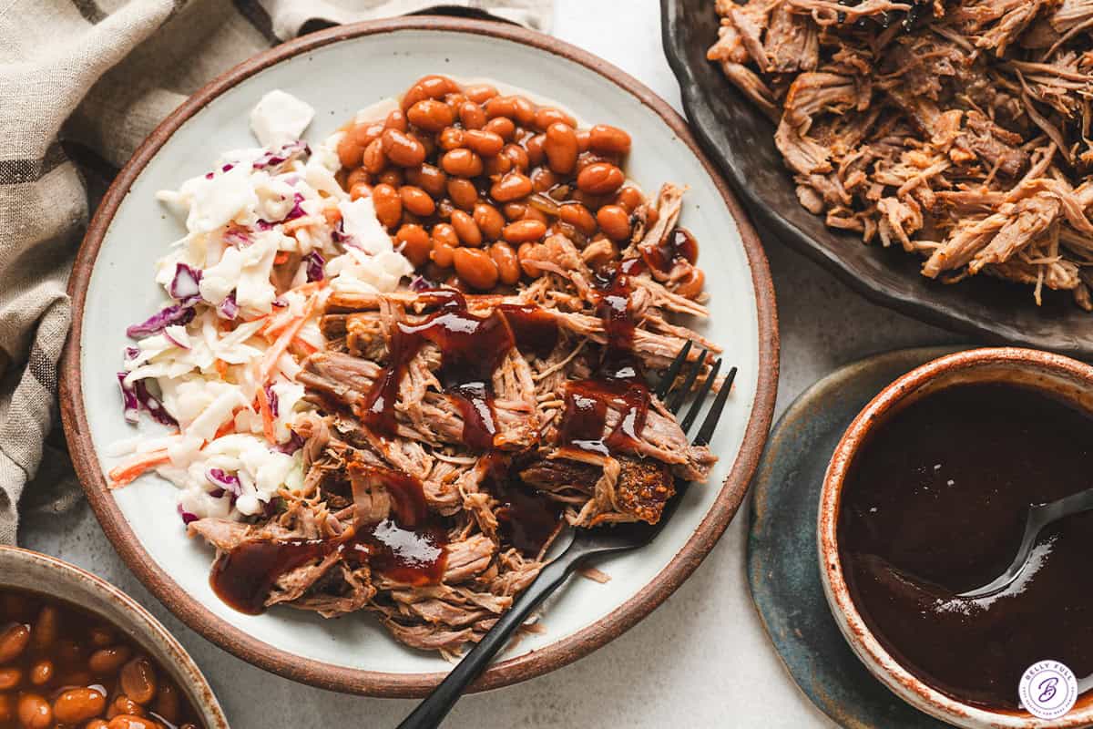 Plate of pulled pork served with baked beans, coleslaw, and barbecue sauce.