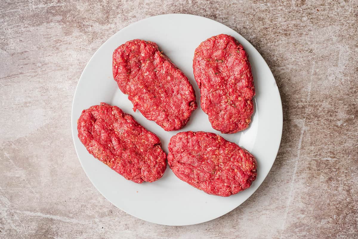 Four raw Salisbury steak patties shaped on a white plate before cooking.