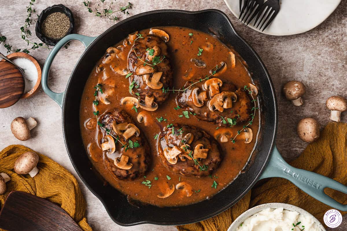 Salisbury steak patties simmered in rich mushroom gravy in a cast iron skillet garnished with fresh herbs.