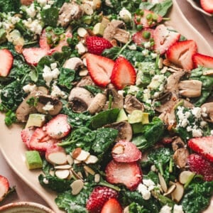 Strawberry spinach salad with fresh strawberries, mushrooms, avocado, goat cheese, sliced almonds, and scallions tossed in creamy poppy seed dressing on a serving platter.