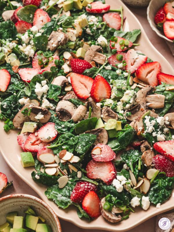 Strawberry spinach salad with fresh strawberries, mushrooms, avocado, goat cheese, sliced almonds, and scallions tossed in creamy poppy seed dressing on a serving platter.