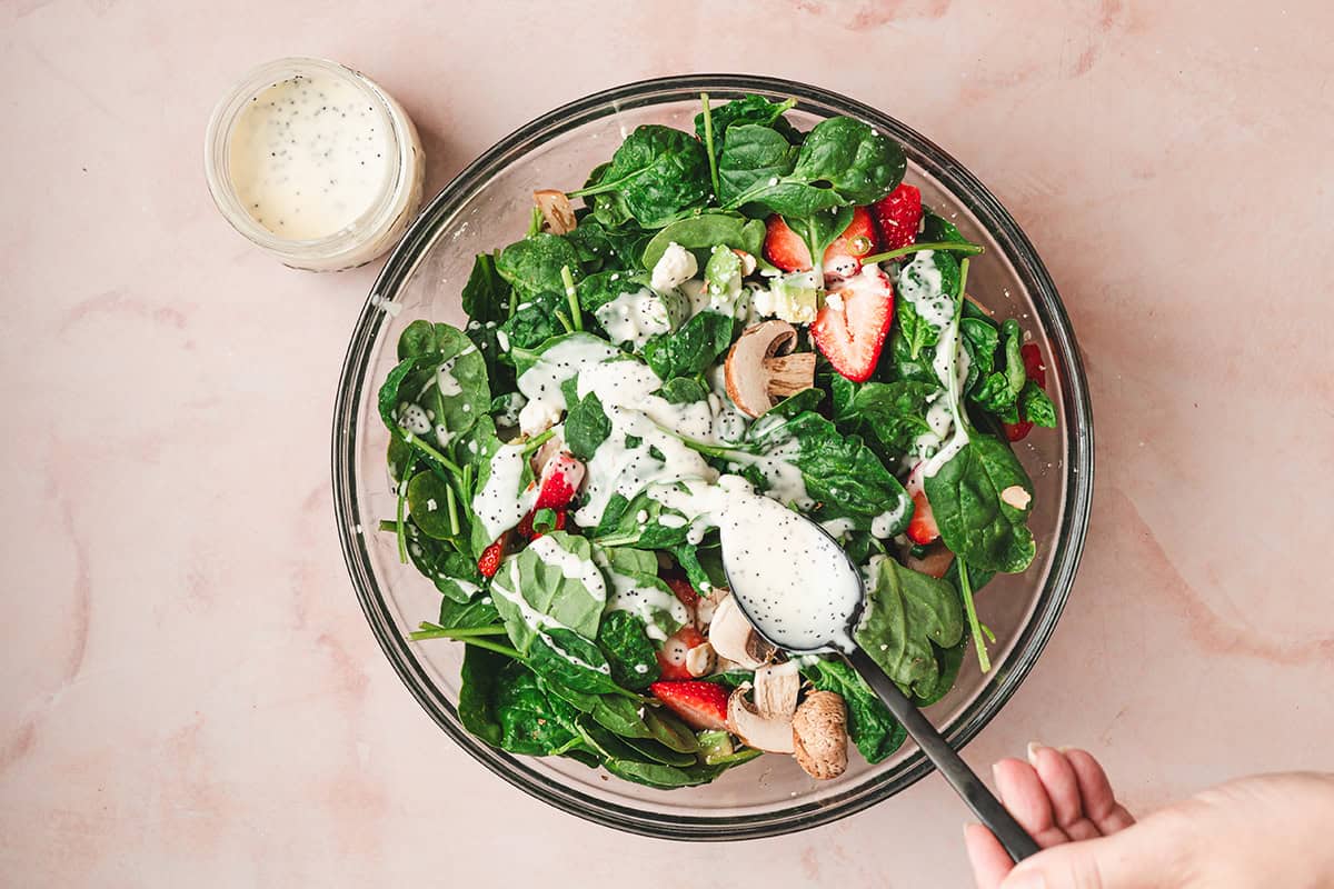 Homemade poppy seed dressing being spooned over a strawberry spinach salad in a large glass bowl.