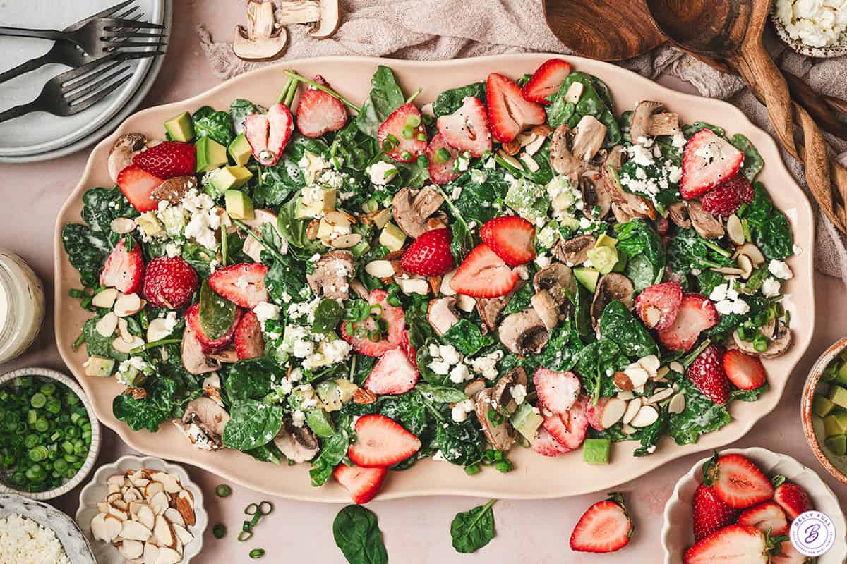 Overhead view of strawberry spinach salad with avocado, mushrooms, goat cheese, sliced almonds, and poppy seed dressing on a serving platter.