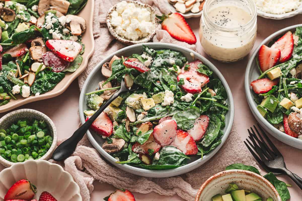 Strawberry spinach salad served in bowls with creamy poppy seed dressing, avocado, mushrooms, goat cheese, and sliced almonds.