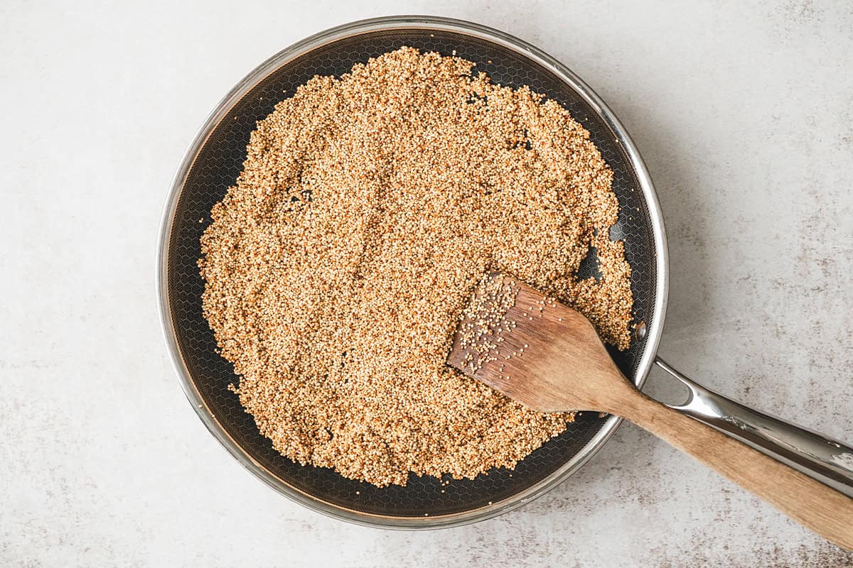 Quinoa toasting in a skillet with olive oil and a wooden spatula.