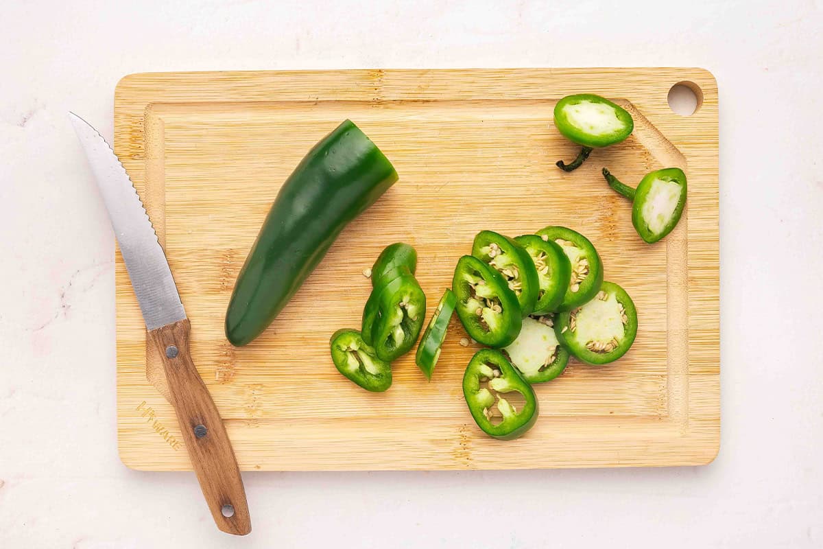 Half a jalapeno on a wooden cutting board with jalapeno slices next to it.