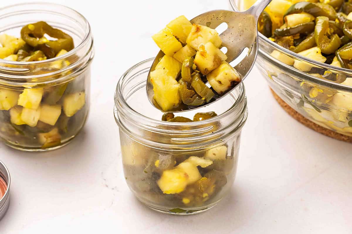 A slotted spoon adding jalapenos and pineapple to a glass mason jar.