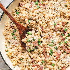 Creamy bacon pea pasta in a skillet topped with grated Parmesan cheese and served with a wooden spoon.