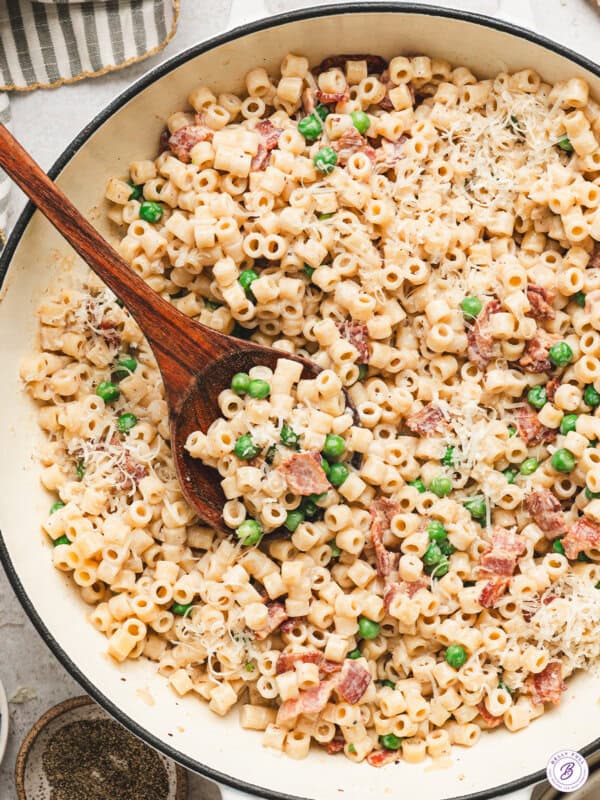 Creamy bacon pea pasta in a skillet topped with grated Parmesan cheese and served with a wooden spoon.
