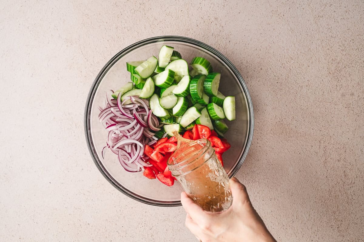 Pouring homemade dressing from a mason jar over chopped cucumbers, tomatoes, and red onion in a glass bowl