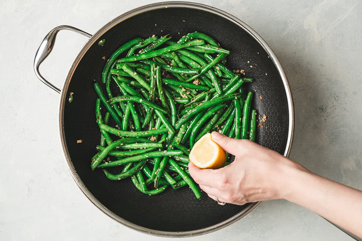 A hand squeezing lemon over garlic green beans in a skillet.