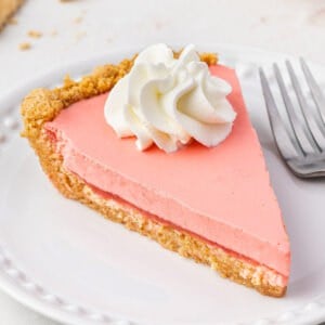 Slice of pink no-bake Jello pie topped with whipped cream on a white dessert plate.