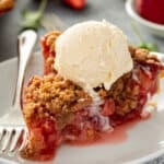Slice of strawberry rhubarb pie topped with a scoop of vanilla ice cream on a white plate.