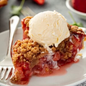 Slice of strawberry rhubarb pie topped with a scoop of vanilla ice cream on a white plate.