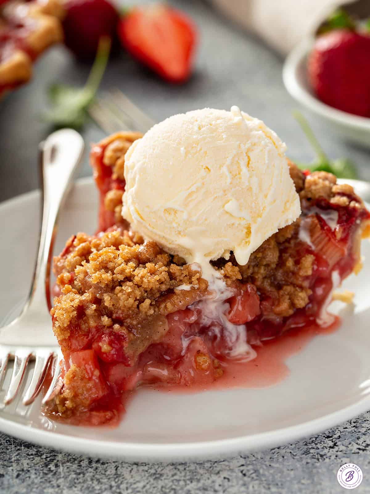 Slice of strawberry rhubarb pie topped with a scoop of vanilla ice cream on a white plate.