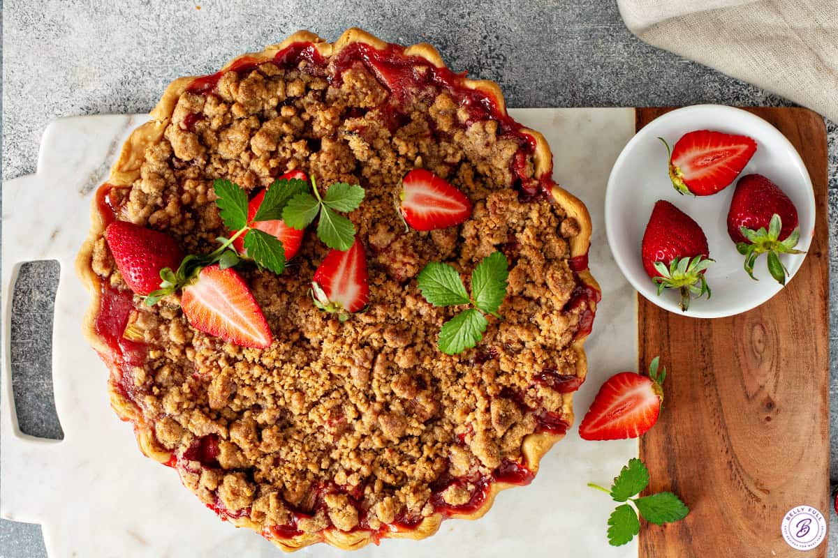 Baked strawberry rhubarb pie with a golden crumb topping garnished with fresh strawberries.