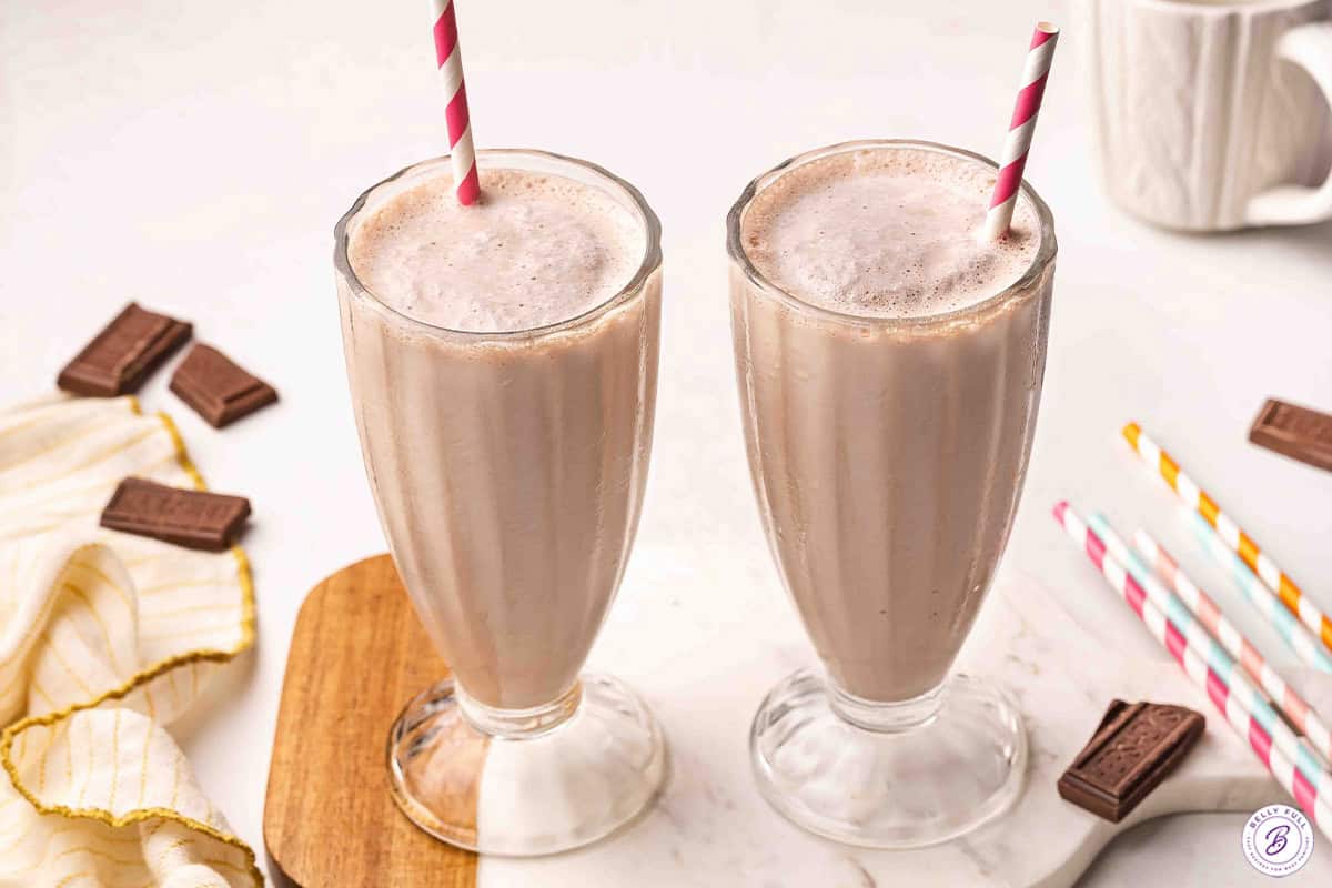 Two tall glasses of homemade Wendy's Frosty copycat with pink striped straws surrounded by chocolate pieces and colorful straws