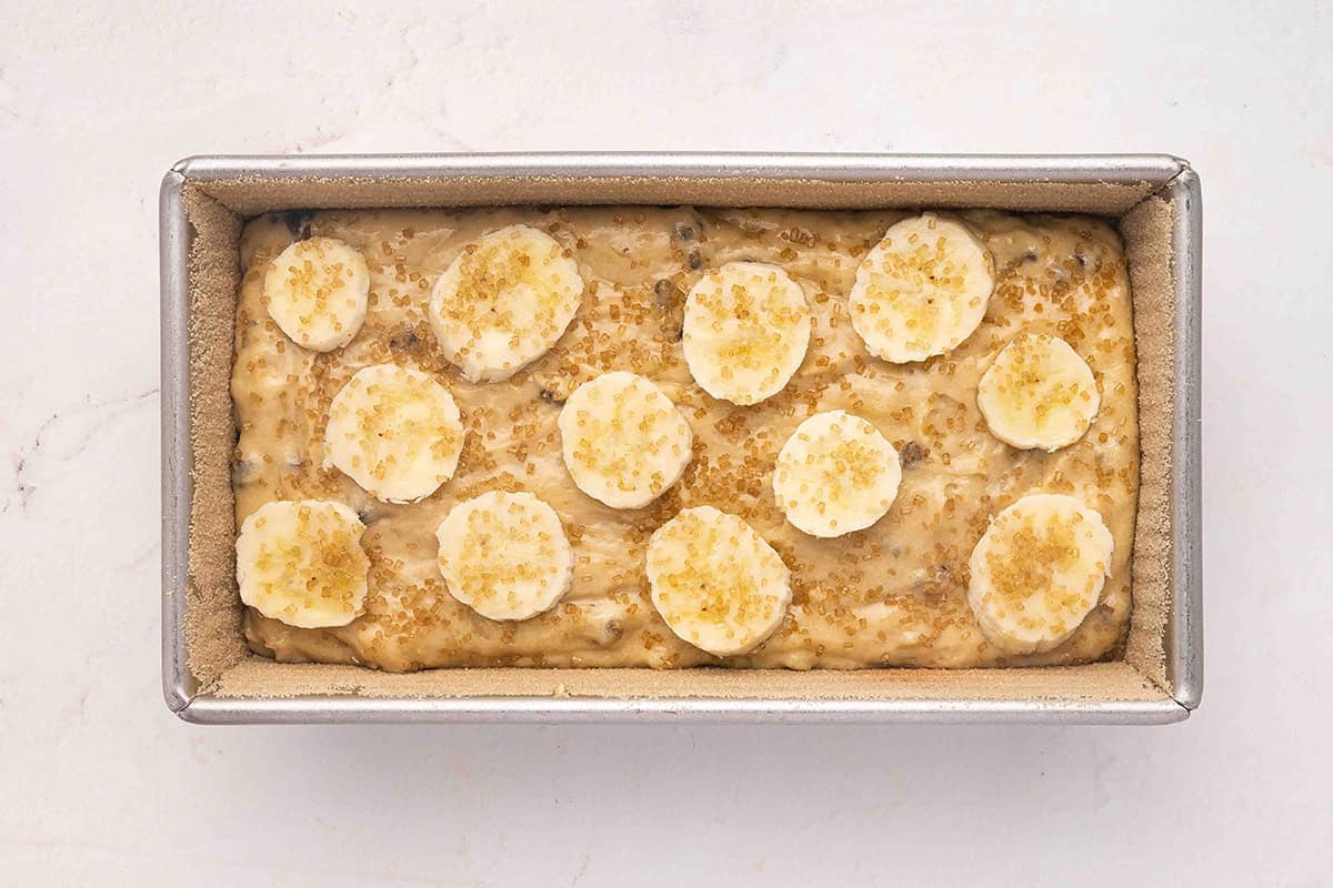 unbaked banana bread batter in loaf pan topped with banana slices and turbinado sugar