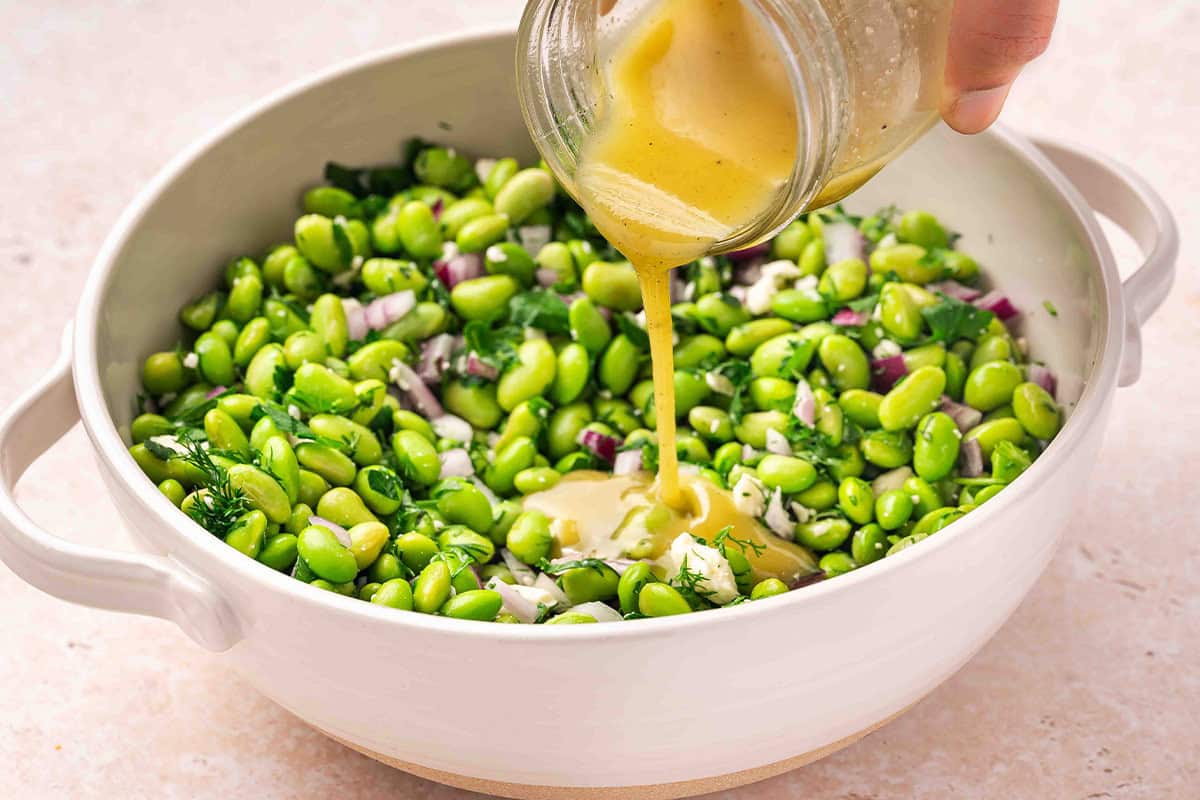Dressing being poured over an edamame salad in a bowl.