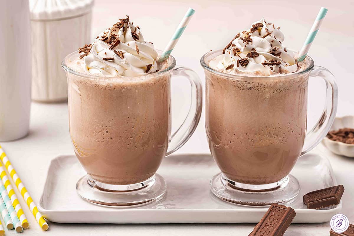 Two mugs of frozen hot chocolate topped with whipped cream and chocolate shavings, served on a tray with chocolate bars
