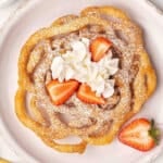 Overhead view of a homemade funnel cake on a plate with whipped cream and strawberries.