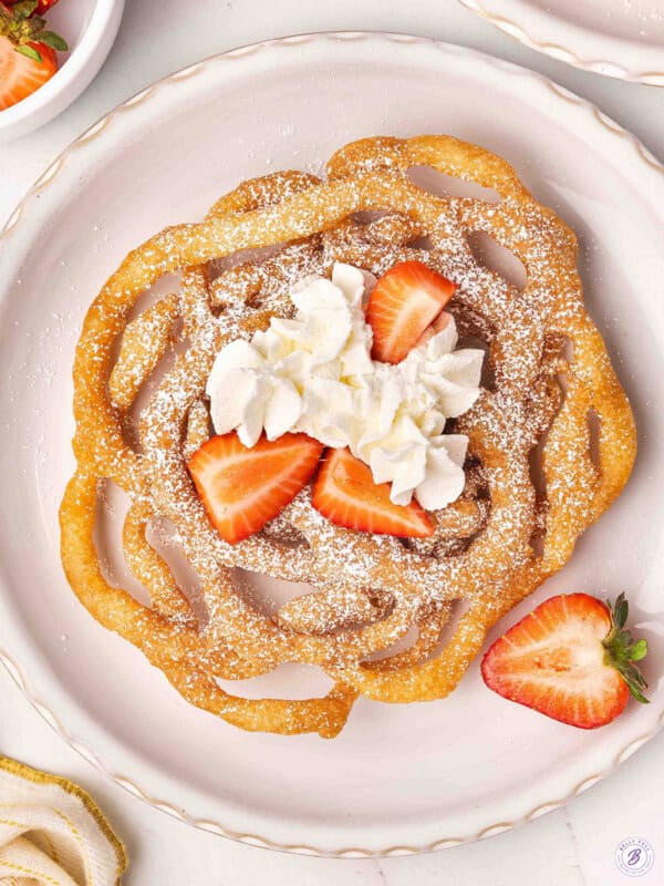 Overhead view of a homemade funnel cake on a plate with whipped cream and strawberries.