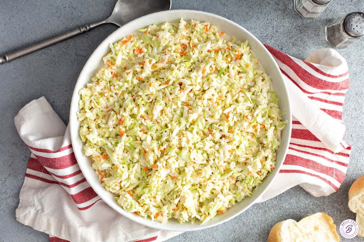 overhead of KFC coleslaw in a large white bowl on a striped napkin with dinner rolls