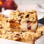 Side view of a peach cobbler cheesecake bar being lifted out of a baking dish.