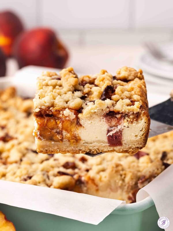 Side view of a peach cobbler cheesecake bar being lifted out of a baking dish.