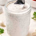 Overhead close-up of homemade ranch dressing in a glass mason jar with a spoon, surrounded by fresh parsley and garlic cloves