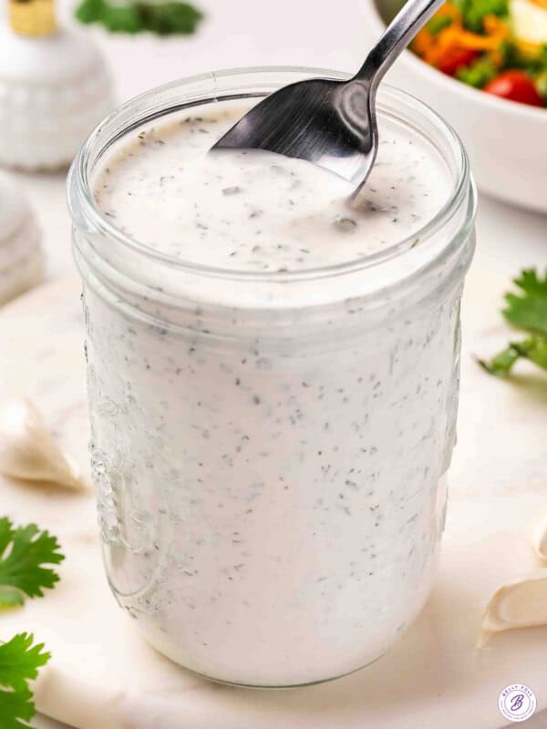 Overhead close-up of homemade ranch dressing in a glass mason jar with a spoon, surrounded by fresh parsley and garlic cloves