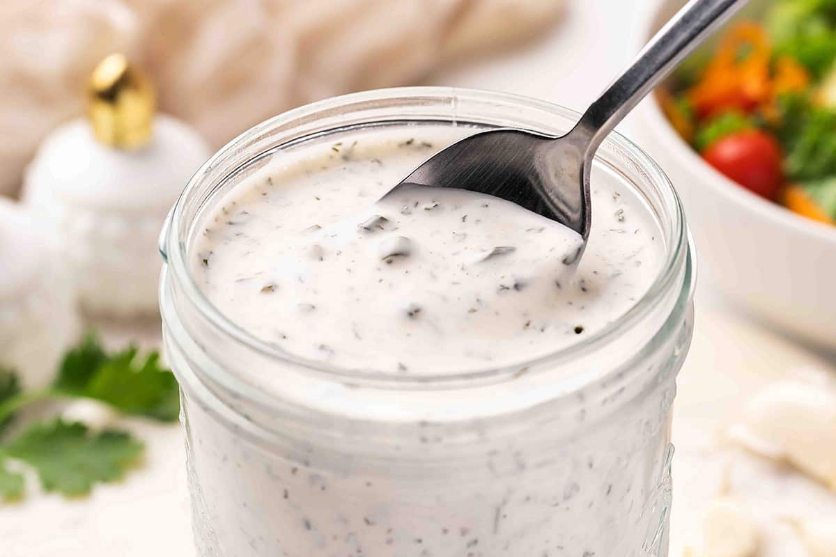 Close-up angle of a spoon stirring creamy homemade ranch dressing in a mason jar, with a fresh salad blurred in the background