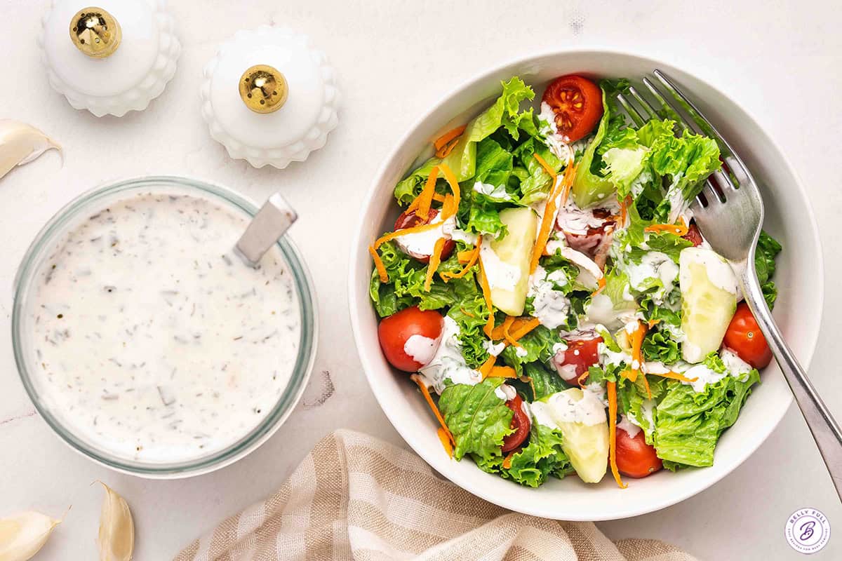 Overhead shot of a bowl of ranch dressing next to a fresh garden salad with lettuce, cherry tomatoes, cucumber, and shredded carrots drizzled with ranch
