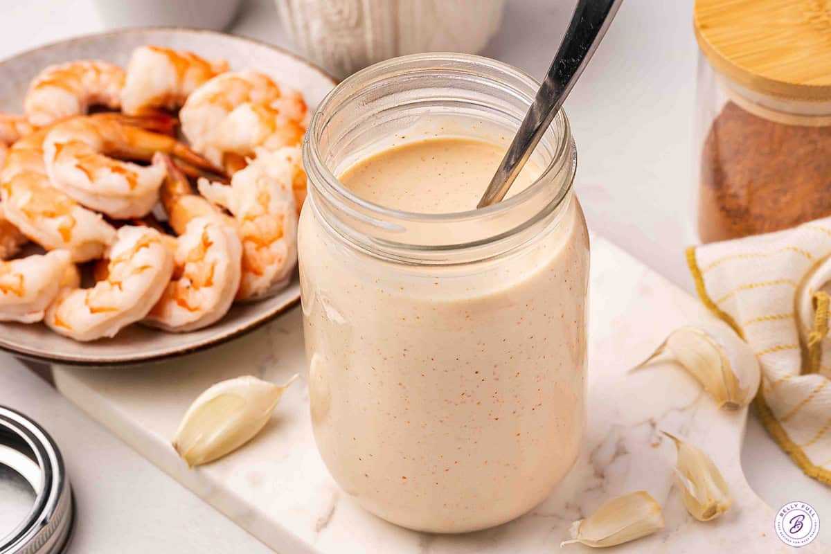 Mason jar of homemade Yum Yum Sauce with a spoon, next to a plate of cooked shrimp