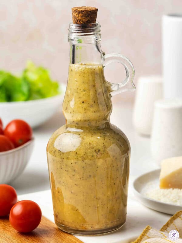 Homemade Italian dressing in a glass cruet bottle with a cork stopper, surrounded by cherry tomatoes and fresh lettuce