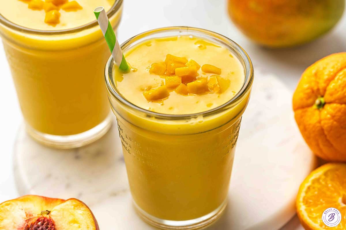 Two glasses of mango orange smoothie topped with diced mango, served with fresh oranges and a peach half.