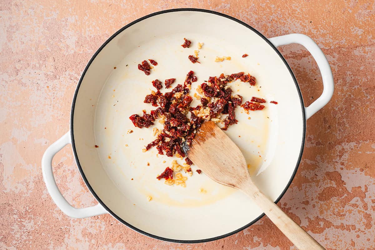 Minced garlic and sun dried tomatoes in a skillet with a wooden spatula.