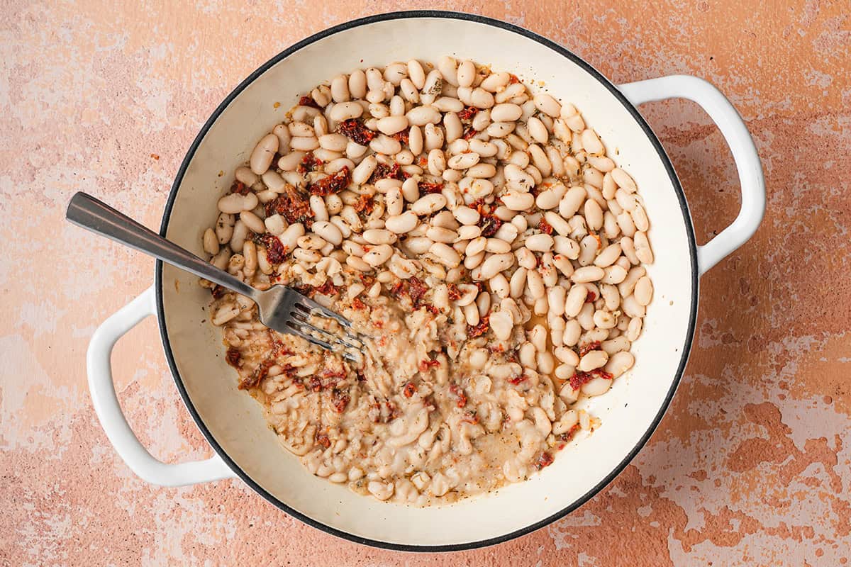 A skillet of white beans and sundried tomatoes, with half mashed by a fork.
