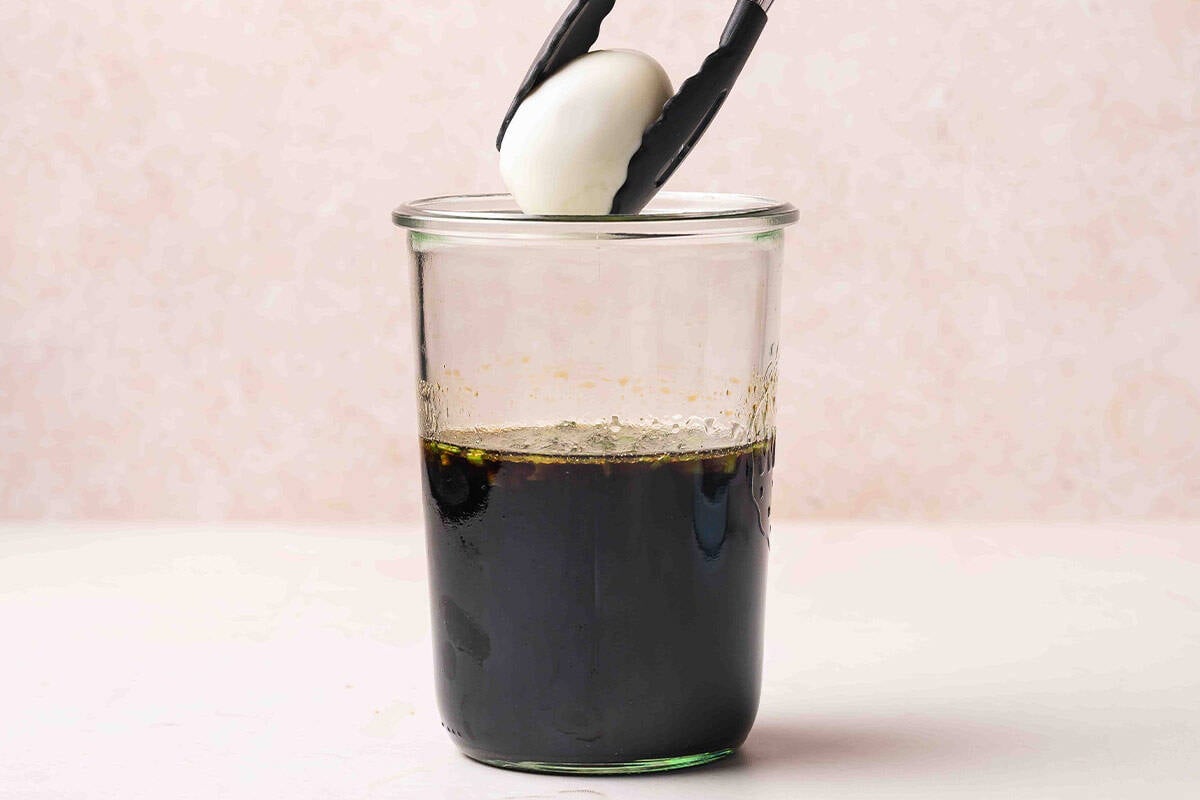 Tongs lowering a peeled hard boiled egg into a jar of soy sauce marinade.