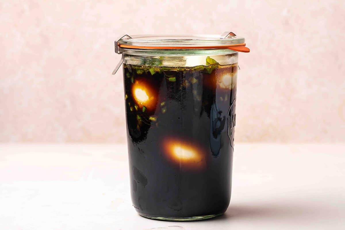 Sealed glass jar of soy sauce eggs marinating in the refrigerator, with scallions visible throughout the dark liquid