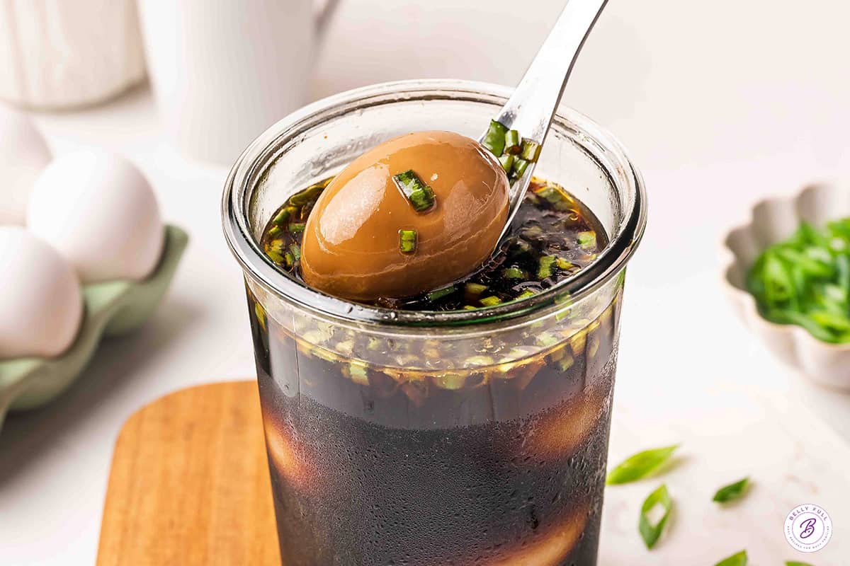 A spoon lifting one finished soy sauce egg out of the jar, coated in marinade and diced scallions