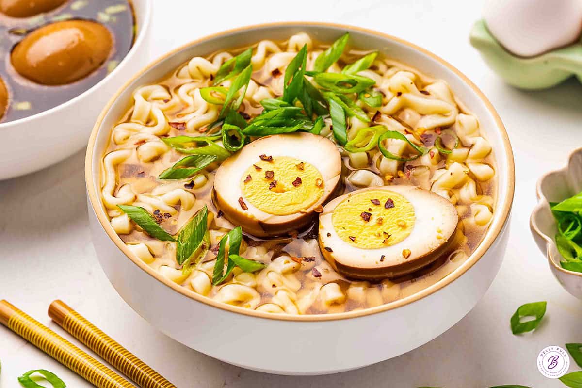 A bowl of ramen noodle soup topped with sliced soy sauce eggs, scallions, and red pepper flakes