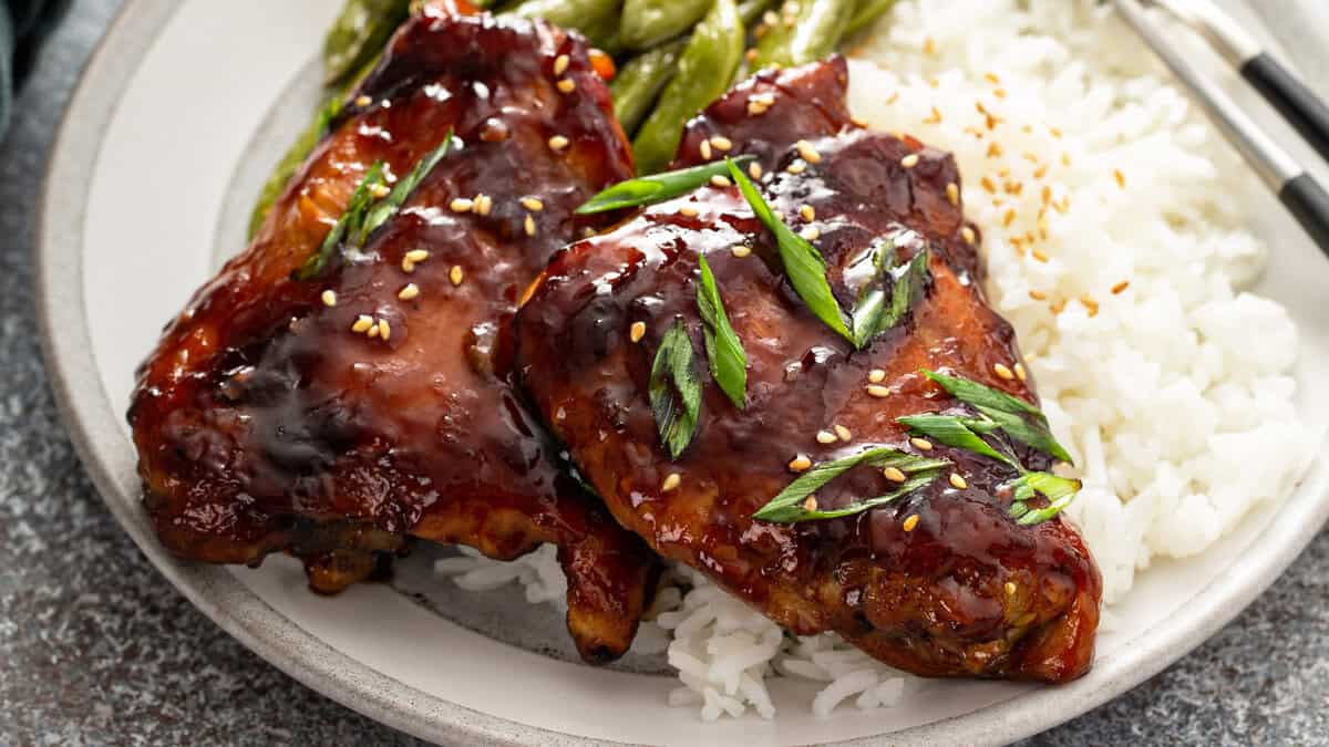 Close-up landscape shot of a plate with two teriyaki chicken thighs over white rice and snap peas, garnished with scallions and sesame seeds.