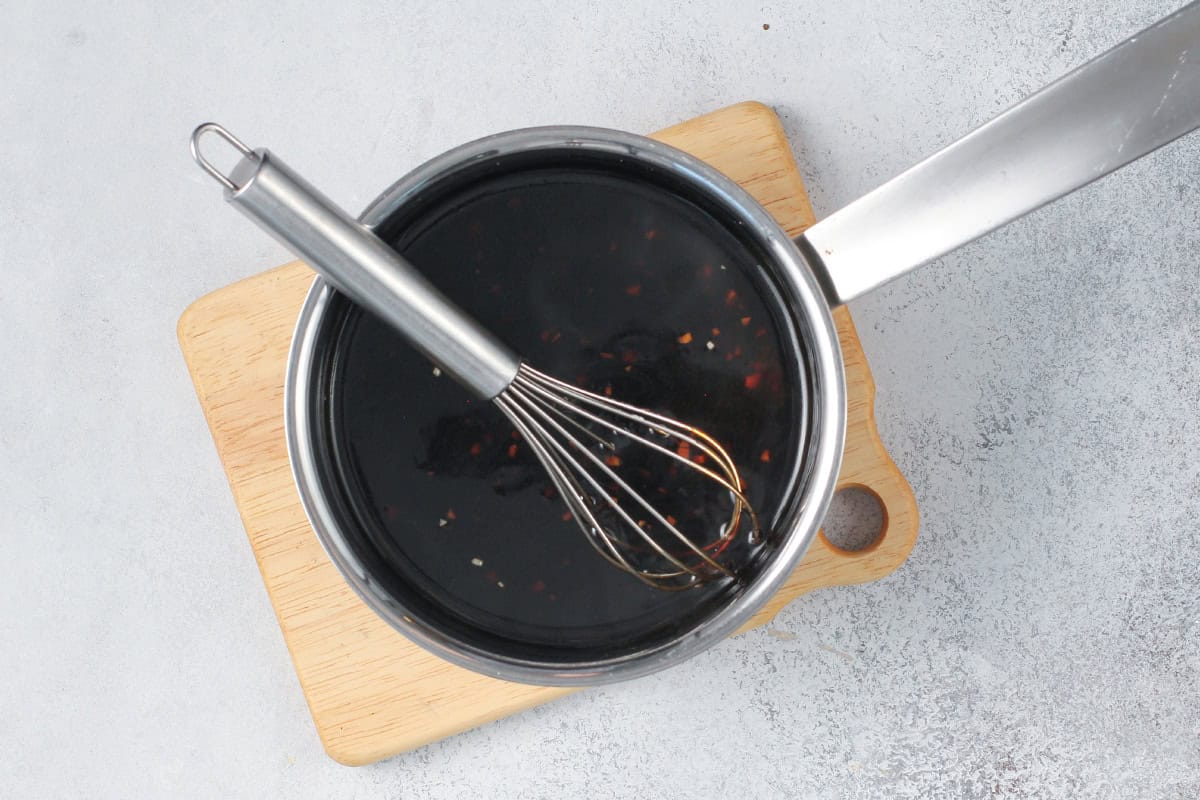 Overhead shot of homemade teriyaki sauce in a small saucepan, dark and glossy with flecks of garlic and ginger, a whisk resting inside.