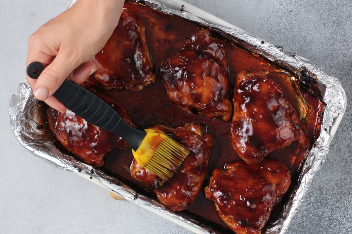 Overhead shot of a hand basting baked teriyaki chicken thighs with a pastry brush, the sauce dark, glossy, and thickened in the foil-lined pan.