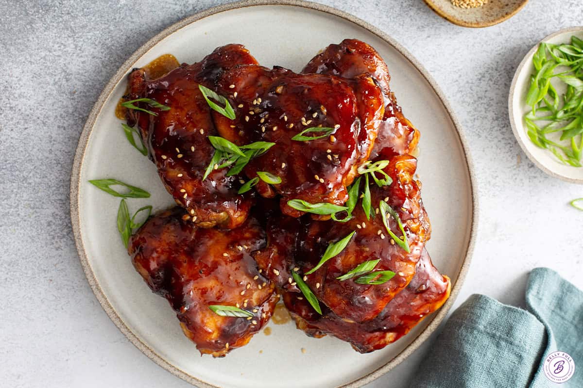 Overhead shot of finished teriyaki chicken thighs on a speckled ceramic plate, garnished with sliced scallions and sesame seeds, with a small bowl of scallions in the background and a blue napkin at the edge of the frame.
